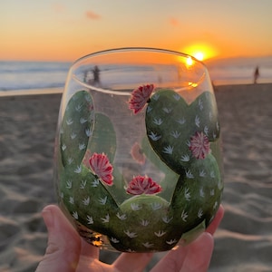 May include: A clear stemless wine glass featuring a hand-painted cactus design. The cactus is green with white accents and red flowers. The glass is held up against a beach sunset.