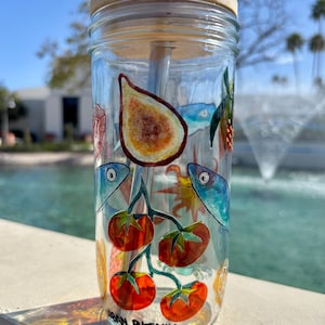 May include: A clear glass tumbler with a bamboo lid and a blue straw. The tumbler is decorated with colorful illustrations of figs, tomatoes, and fish. The text "Urban Phenix" is printed on the bottom of the glass.