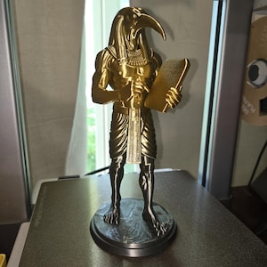 Peut inclure: Une figurine dorée métallique représentant la divinité égyptienne Thot, dotée d'une tête d'ibis et d'un corps humain. La figure porte un pagne et un collier traditionnels, tenant une tablette rectangulaire sur un socle circulaire.
