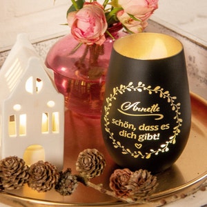 Könnte beinhalten: Schwarzer Glas-Kerzenhalter mit goldfarbenem Rand und einer goldenen Inschrift mit dem Text "Annette schön, dass es dich gibt!" mit einem kleinen Herzsymbol. Der Kerzenhalter ist von Tannenzapfen und einer Vase mit rosa Rosen umgeben.