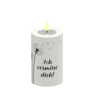 Op de afbeelding: Een witte cilindrische kaars met een paardebloem ontwerp en de tekst "Ich vermisse dich!" in het zwart. De kaars heeft een gele vlam en een witte rand.