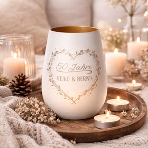 Op de afbeelding: Een witte keramische kandelaar met een gouden rand en een hartvormig ontwerp. Het ontwerp bevat de tekst "50 Jahre HEIKE & BERND". De kandelaar is omgeven door brandende kaarsen en decoratieve elementen.