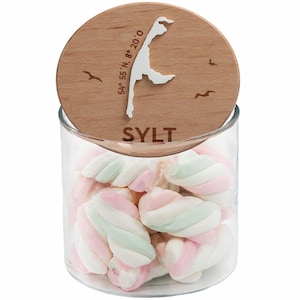 Op de afbeelding: Een glazen pot gevuld met pastelkleurige marshmallows, bekroond met een houten deksel. Het deksel heeft een witte omtrek van een eiland, het woord "SYLT" en coördinaten. De marshmallows zijn gewerveld met roze, wit en groen.