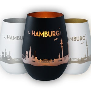 Op de afbeelding: Drie kaarshouders met een stadsbeeld-silhouetontwerp. De kaarshouders zijn in zwart, wit en goud. Het stadsbeeld-silhouet is van Hamburg, Duitsland. De tekst "HAMBURG" is afgedrukt op de voorkant van elke kaarshouder.