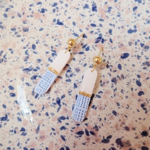 Op de afbeelding: Een paar hangende oorbellen met een goudkleurige bolvormige knop bevestigd aan een lange, afgeronde rechthoekige hanger. De hanger is verdeeld in een effen crèmekleurige bovenkant en een lichtblauw ondergedeelte met een rasterpatroon.