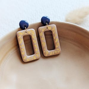 Könnte beinhalten: Ein Paar rechteckige Hängeohrringe mit goldfarbenem Glitzerdesign und marineblauen Akzenten.