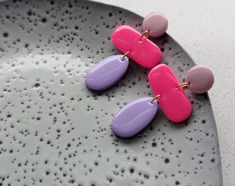 Könnte beinhalten: Ein Paar lebendige Ohrringe mit rosa, lila und hellrosa Elementen. Die Ohrringe haben einen goldfarbenen Haken und bestehen aus einem kleinen hellrosa Kreis, einer rosa länglichen Form und einer lila ovalen Form. Die Ohrringe werden auf einem grau gesprenkelten Teller präsentiert.