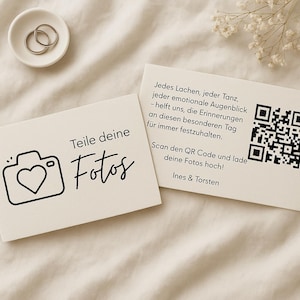 Könnte beinhalten: Zwei cremefarbene rechteckige Karten mit schwarzem Text und Grafiken. Eine Karte zeigt ein Kamerasymbol mit Herz und den Text "Teile deine Fotos". Die andere Karte enthält einen QR-Code und deutschen Text zum Hochladen von Fotos für einen Tag.