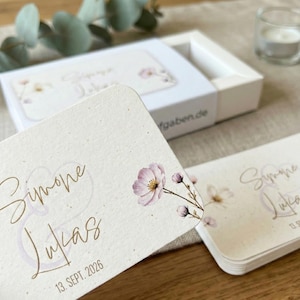 Puede incluir: Tarjetas color crema con esquinas redondeadas, con nombres en escritura dorada y la fecha "13. Sept. 2026." Un delicado diseño floral en rosa y morado adorna la esquina. Las tarjetas están en una caja blanca.