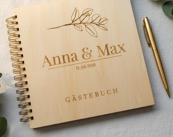 May include: A light-colored wooden guest book with a spiral binding. The cover is engraved with the names "Anna & Max", the date "12. Juli 2026", and the word "GÄSTEBUCH". A gold pen rests beside the book.