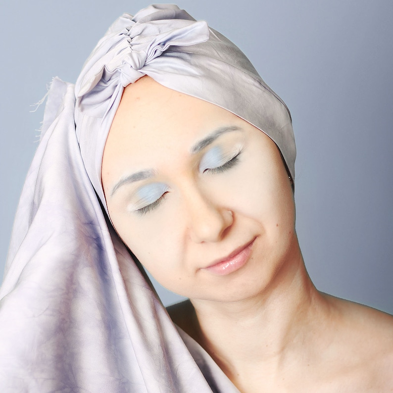 May include: A woman with her eyes closed wearing a light purple turban. Her face is lightly powdered and her eyelids are highlighted with a light blue eyeshadow.