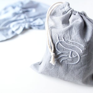 May include: A gray drawstring bag with a white drawstring and a white embroidered design on the front. The design is a stylized letter "S" with a face.