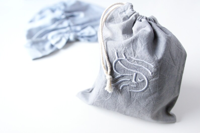 May include: A gray drawstring bag with a white drawstring and a white embroidered design on the front. The design is a stylized letter "S" with a face.