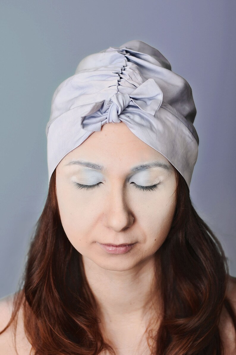 May include: A woman with long brown hair wears a light blue turban with a bow on top. Her eyes are closed and her face is covered in white powder.