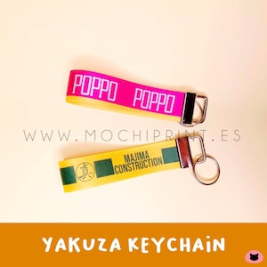 Könnte beinhalten: Zwei Yakuza-Schlüsselanhänger. Einer ist pink mit dem Text "POPO", der andere ist gelb mit dem Text "MAJIMA CONSTRUCTION". Beide haben silberne Schlüsselringe und schwarze Metallverschlüsse. Die Website www.mochiprint.es ist sichtbar.