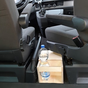 Op de afbeelding: Een houten opbergplank met twee compartimenten is geïnstalleerd in het passagiersgebied van een voertuig. Een doorzichtige plastic waterfles staat op de plank.