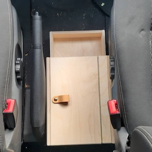 May include: A wooden storage box with a hinged lid and a leather handle. The box is installed in the center console of a car.