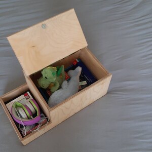 May include: A wooden box with a hinged lid, open to reveal a green and gray stuffed animal, a book, and a pair of purple headphones in a separate compartment.