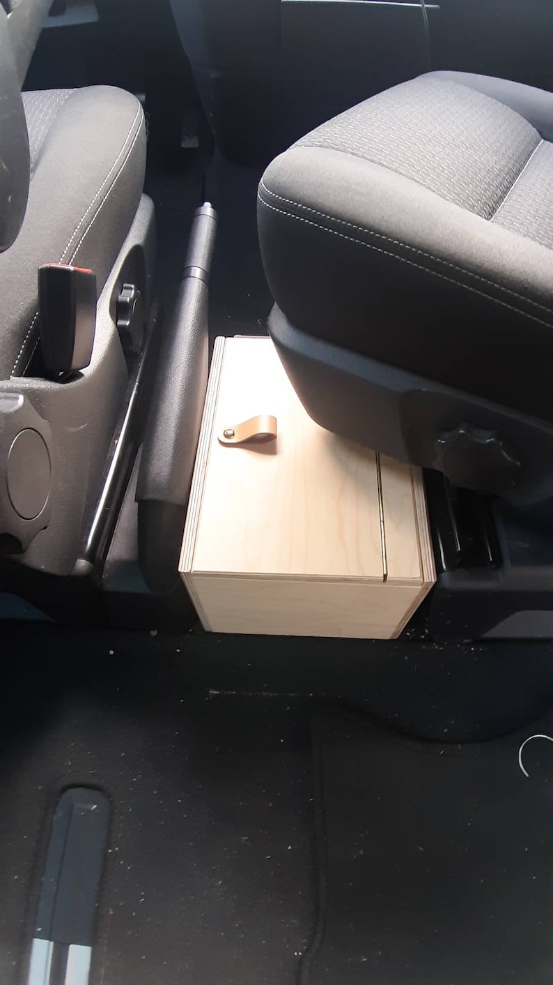 May include: A light wood storage box with a leather handle, installed between the front seats of a car.
