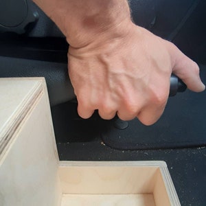 May include: A person's hand pulling a black handbrake lever in a car. The handbrake is attached to a wooden box.