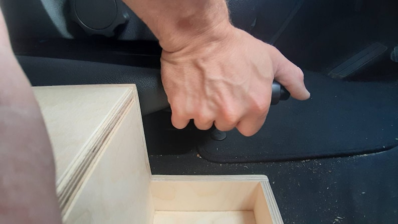 May include: A person's hand pulling a black handbrake lever in a car. The handbrake is attached to a wooden box.
