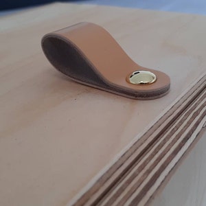 May include: A wooden box with a light brown leather handle and a gold metal accent. The box is made of plywood and has a natural wood finish.