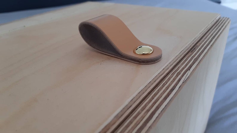 May include: A wooden box with a light brown leather handle and a gold metal accent. The box is made of plywood and has a natural wood finish.