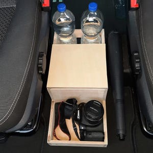 May include: A wooden storage box with a lid, designed to fit between two car seats. The box has two compartments for holding water bottles and a lower compartment for storing a camera. The box is made of light wood and has a natural finish.