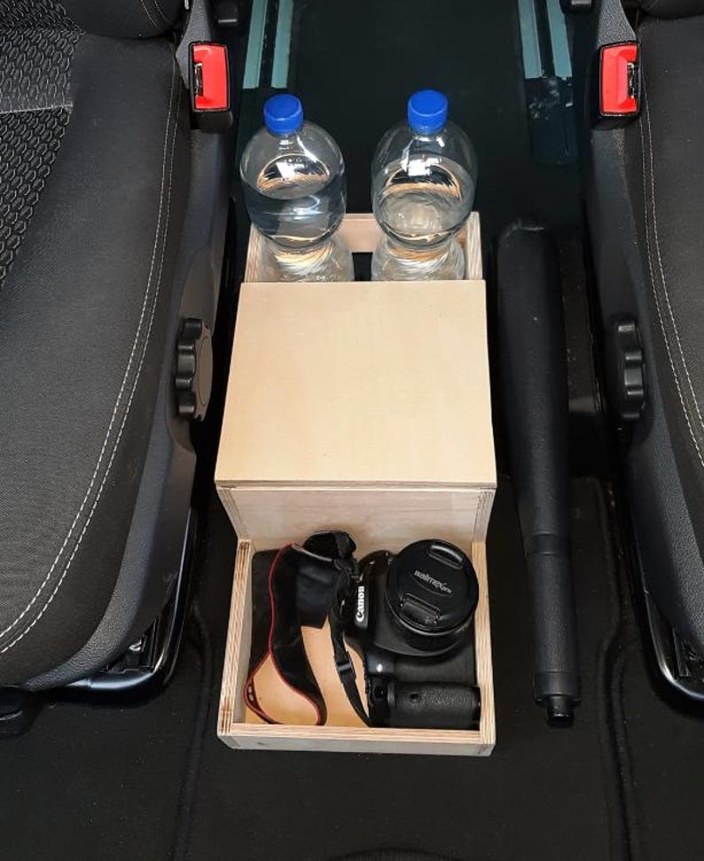 May include: A wooden storage box with a lid, designed to fit between two car seats. The box has two compartments for holding water bottles and a lower compartment for storing a camera. The box is made of light wood and has a natural finish.