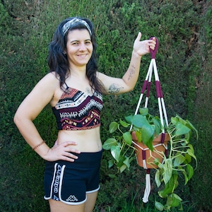 Puede incluir: Una maceta de terracota con hojas verdes, suspendida por un colgador de plantas de macramé. El colgador tiene cuerdas blancas y burdeos. La maceta está sostenida por una persona.