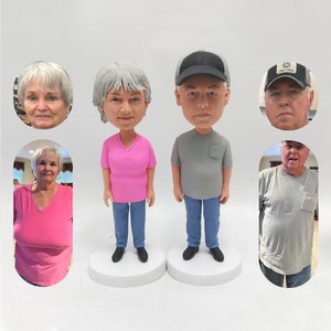 May include: Two custom-made bobbleheads, one with gray hair and a pink shirt, the other with gray hair and a gray shirt. Both bobbleheads are wearing blue jeans.