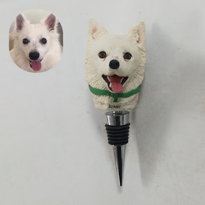 Puede incluir: Un tapón de botella con forma de perro blanco con un collar verde y el nombre "Benny" grabado. El tapón tiene una base de metal plateado y un sello de goma negro. Una imagen más pequeña del perro está en la esquina superior izquierda.