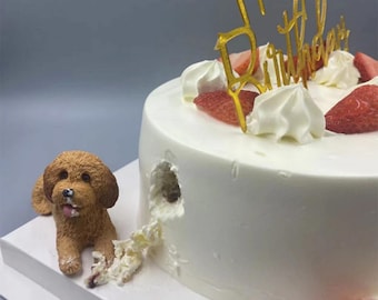 Puede incluir: Un pastel blanco con un adorno dorado de "Feliz cumpleaños", fresas y crema batida. Una pequeña figurita de perro marrón está dando un mordisco al pastel.