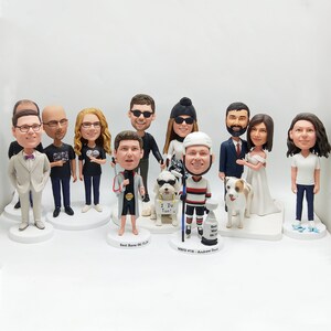 May include: A collection of custom bobblehead figurines, each depicting a person or couple. The figures vary in attire, including formal wear, casual outfits, and sports uniforms. Some figurines are accompanied by small dog figures. Each bobblehead is on a white base.