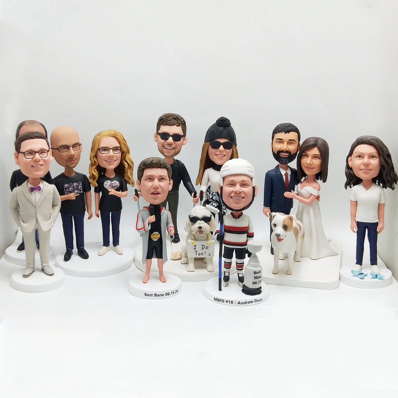 May include: A collection of custom bobblehead figurines, each depicting a person or couple. The figures vary in attire, including formal wear, casual outfits, and sports uniforms. Some figurines are accompanied by small dog figures. Each bobblehead is on a white base.