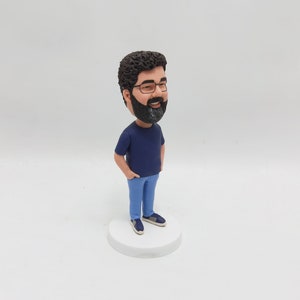 May include: A custom-made figurine of a person with brown hair and a beard wearing a blue shirt and blue jeans. The figurine is standing on a white base.