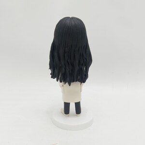 May include: A handmade figurine of a person with long, black hair, wearing a white coat and black pants. The figure is standing on a white circular base. The back of the figure is facing the viewer.