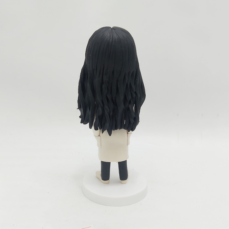 May include: A handmade figurine of a person with long, black hair, wearing a white coat and black pants. The figure is standing on a white circular base. The back of the figure is facing the viewer.