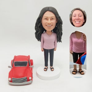 May include: A custom figurine of a person with long dark hair wearing a pink shirt and black pants. The figurine is standing on a white base next to a red toy car. The person's face is smiling and their eyes are brown. The figurine is a personalized gift.