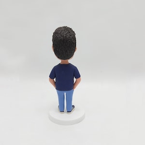 May include: A blue and white figurine of a person with short brown hair. The figurine is standing with hands in pockets and is wearing a blue shirt and blue jeans.