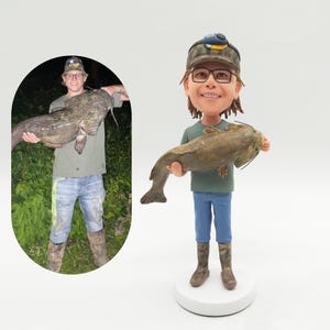 May include: A custom figurine of a person holding a large catfish. The figurine is wearing a green shirt, blue jeans, and camouflage boots. The person is wearing a camouflage hat and glasses. The figurine is standing on a white base.