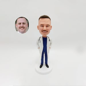 Könnte beinhalten: Eine personalisierte Bobblehead-Figur eines Arztes, der einen weißen Kittel und blaue Scrubs trägt. Die Figur hat ein Stethoskop um den Hals und steht auf einem weißen Sockel.