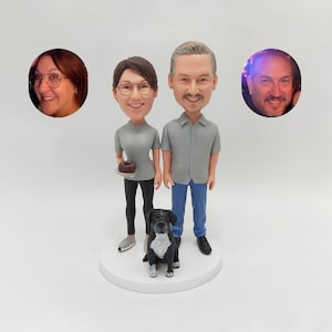 May include: A custom-made figurine set of a couple with a black dog. The couple are wearing grey shirts and jeans. The woman is holding a small cake. The figurines are standing on a white base.
