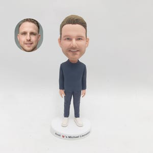 May include: A custom bobblehead figurine of a person with brown hair wearing a dark blue shirt and dark blue pants. The figurine is standing on a white base with the text "Knot's Michael Lisovetsky" written on it.