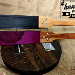 May include: Two leather paddles with wooden handles. The top paddle is black leather and the bottom paddle is purple leather with a decorative design. Both paddles have metal rivets on the handle. The text "hand made in GERMANY" is visible in the top right corner of the image.