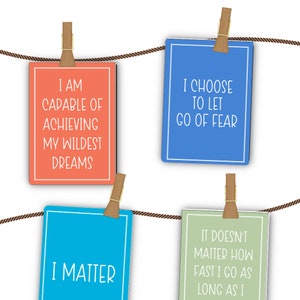 May include: Four colorful cards with inspirational quotes hanging on a clothesline. The quotes are: "I am capable of achieving my wildest dreams", "I choose to let go of fear", "I matter", and "It doesn't matter how fast I go as long as I".