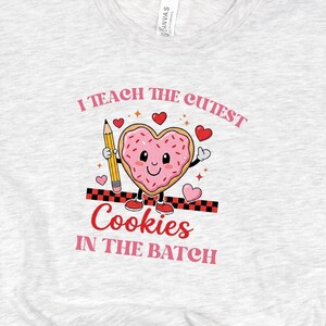 May include: A light gray t-shirt with the text "I teach the cutest cookies in the batch." The graphic features a heart-shaped cookie with a smiling face, holding a pencil. The shirt is paired with black jeans and leopard print flats.