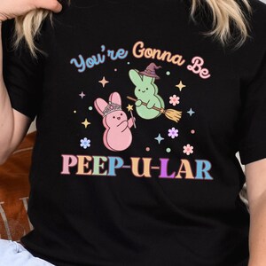 May include: Black t-shirt with the text "You're Gonna Be PEEP-U-LAR" in colorful lettering. The graphic features two cartoon marshmallow bunnies, one pink with a crown and the other green with a witch hat, on a broom.