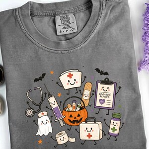 May include: A charcoal gray crew neck t-shirt featuring a graphic print of cartoon medical supplies and a ghost with smiling faces. The design includes a stethoscope, syringe, bandage, clipboard labeled BOO-BOO REPORT, pumpkin bucket, and first aid kit.