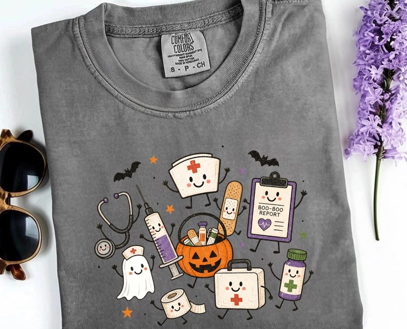 May include: A charcoal gray crew neck t-shirt featuring a graphic print of cartoon medical supplies and a ghost with smiling faces. The design includes a stethoscope, syringe, bandage, clipboard labeled BOO-BOO REPORT, pumpkin bucket, and first aid kit.